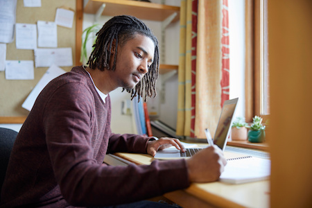 young man working at his desk at home lookin gat his laptop while he takes notes - best free online colleges in Louisiana
